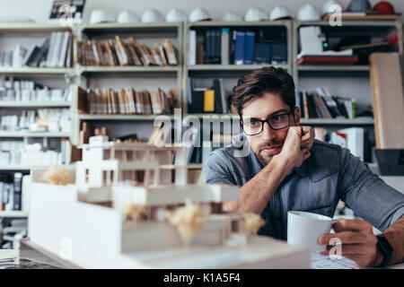 Thoughtful male designer in office looking at model with cup of coffee. Young male architect taking break from work. Stock Photo