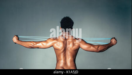 Rear view of muscular young man stretching arms with rubber band. Male bodybuilder doing exercises with elastic band on grey background. Stock Photo