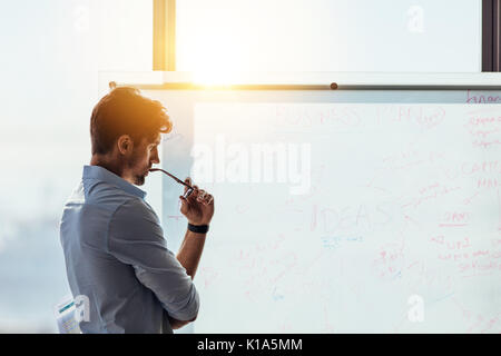 Business investor in deep thought looking at the business ideas written on the whiteboard. Businessman thinking while holding his spectacles to mouth Stock Photo