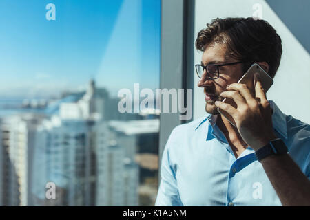 Young businessman speaking on mobile phone looking away in office. businessman standing beside window of highrise office building. Stock Photo