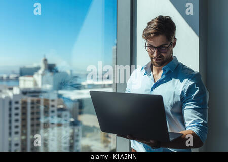 Young businessman working on a laptop computer standing in office. Man holding laptop standing near window of highrise office building overlooking the Stock Photo