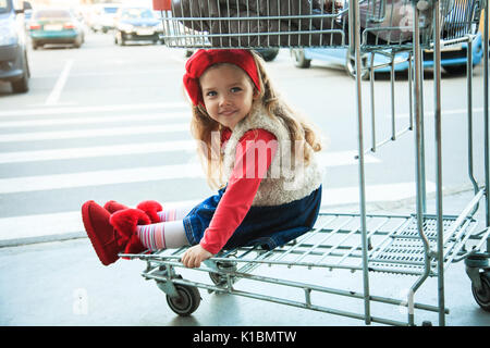 A little sweet girl is sitting in a supermarket shopping cart ootdoors. Stock Photo