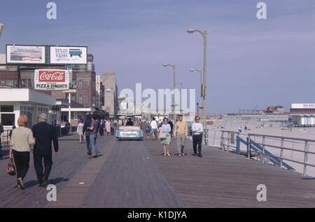 Tourists strolling on the boardwalk in Atlantic City, New Jersey, with signs advertising Coca Cola, a computer printer, and a pizza restaurant, beach visible, 1975. Stock Photo