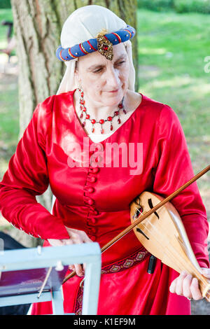 England, Sandwich. Living History player, woman, in red medieval costume, standing under a tree playing a rebec, the fore-runner to the violin. Stock Photo