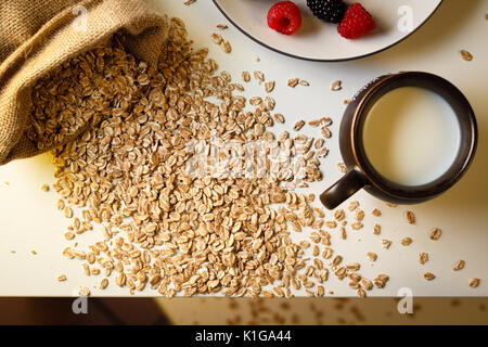 Mess breakfast: scattered oat cereal, milk in cup, blackberry and raspberry on plate, top view. Stock Photo
