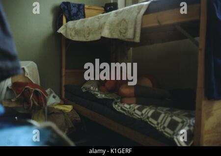 Soft image of backpacker wearing knee socks, curled up and sleeping on the lower bunk of a bunk bed in a youth hostel, bags and other items piled on the floor, Scotland, 1966. Stock Photo