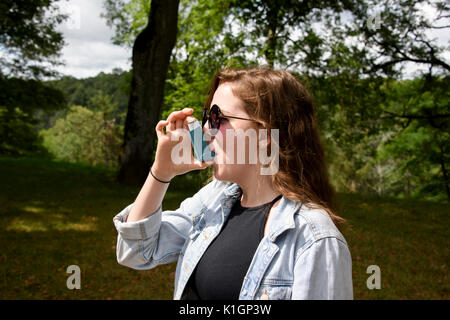 Long haired, teenage girl in denim jacket using a blue asthma inhaler. Taken in woodland on a sunny day. Stock Photo