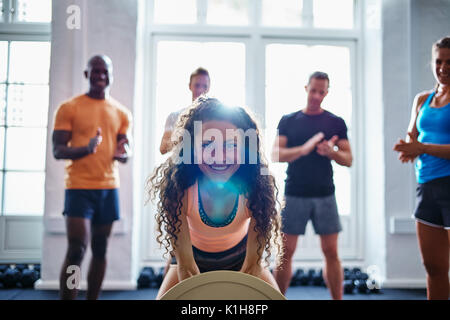 Smiling young woman weightlifting in a gym with a diverse group of friends cheering her on in the background Stock Photo