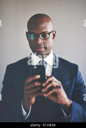 Confident young African businessman wearing a suit and glasses reading text messages on a cellphone while standing alone in an office Stock Photo