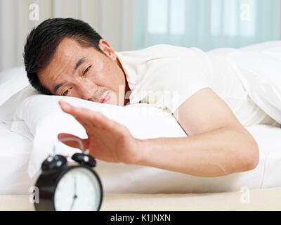 asian man lying in bed trying to stop ringing alarm clock. Stock Photo