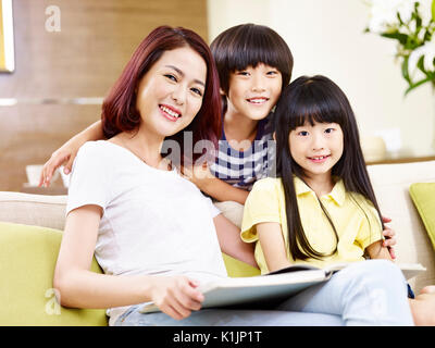 asian mother and two children sitting on couch reading a book. Stock Photo