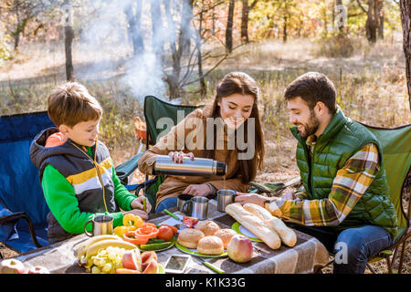 Happy smiling family drinking tea while sitting at table on picnic in autumn forest Stock Photo