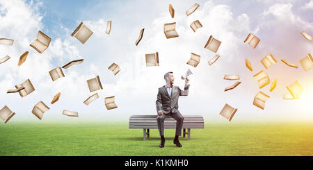 Young businessman sitting on bench and screaming emotionally in megaphone Stock Photo