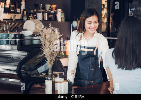 Asian woman barista wear jean apron holding coffee cup served to customer at bar counter with smile emotion,Cafe restaurant service concept,Owner smal Stock Photo