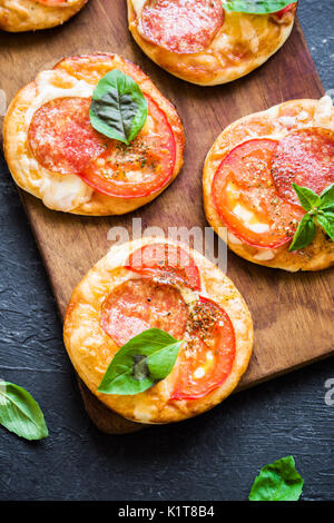 Mini Pizza - Fresh homemade mini pizzas with pepperoni, cheese, tomatoes and basil on rustic black stone background. Stock Photo