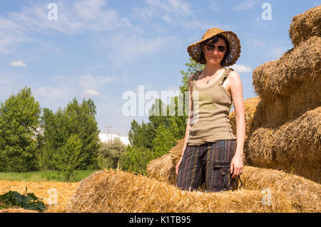 Pretty young woman with hat standing in straw bales and looking to the camera. Stock Photo