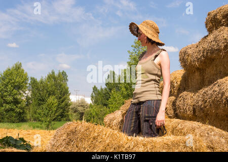 Pretty young woman with hat standing in straw bales and looking away on a sunny day. Stock Photo
