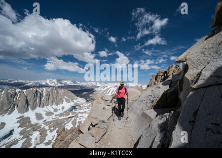 A woman hikes along Trail Crest, on the way down from the summit of Mt. Whitney, overlooking eastern peaks of Sequoia National Park, covered in snow. Stock Photo