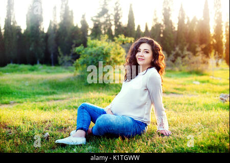 Portrait of happy young pregnant model sitting with crossed legs on grass lawn and looking at her belly with gentle smile. Future mom expecting baby caressing her tummy. Copy space Stock Photo