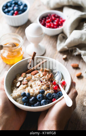 Oatmeal porridge bowl with superfoods in hands. Hands holding bowl of oat porridge with blueberries, almonds, linseed, cinnamon and currants. Selectiv Stock Photo
