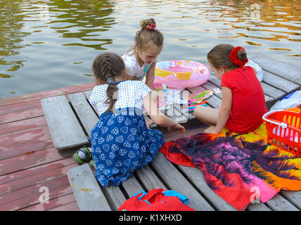 Three little girls play with toys on the wooden floor near the pond. August 2017 Stock Photo