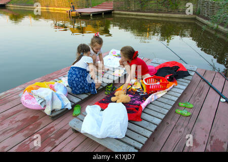 Three little girls play with toys on the wooden floor near the pond. August 2017 Stock Photo