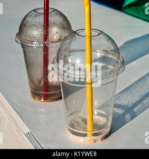 Empty plastic glasses with red and yellow straws stand on a white deck. Some green cloth and reflection of blue object in the background. Sunlit abstr Stock Photo