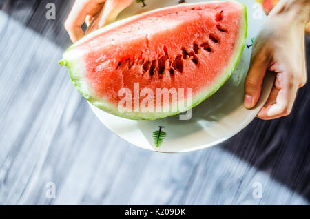 Slice of watermelon on white plate in woman hands ready for eating, top view with blurry black wood table background Stock Photo