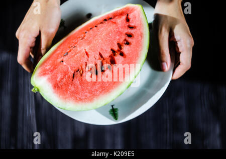 Slice of watermelon on white plate in woman hands ready for eating, top view with blurry black wood table background Stock Photo