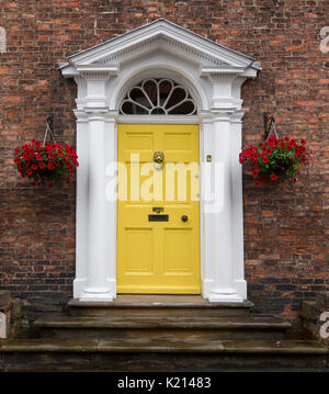 Yellow painted wooden door with white classic style frame / portico bordered by hanging baskets of bright red flowers against dark brick wall Stock Photo