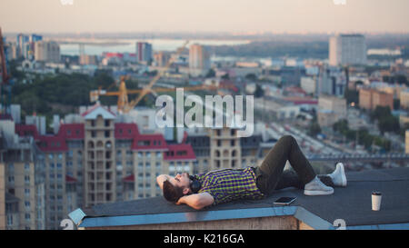 A hipster guy listens to music on the roof of a high-rise building in the city center. He relaxes after a day's work and lies on the roof. Stock Photo