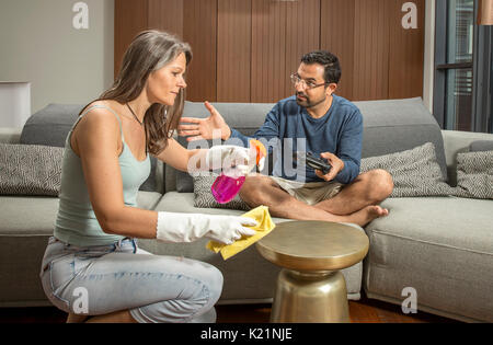 arab man playing a video game while woman is cleaning around him in their city apartment Stock Photo