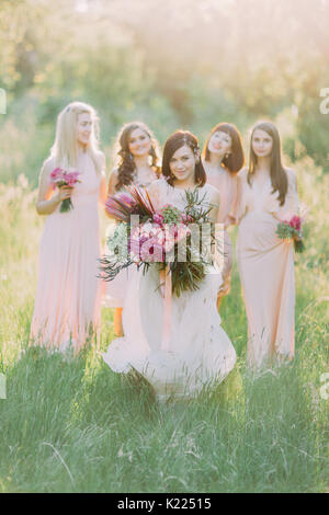 The bride in the long white dress is holding the huge bouquet and going forward at the background of the standing bridesmaids in the spring. Stock Photo