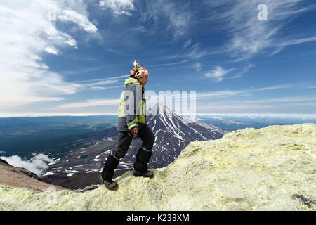 Young women tourists climb to sulfur fumarole field in crater of active Avachinsky Volcano on background of cone of Koryak Volcano on Kamchatka. Stock Photo