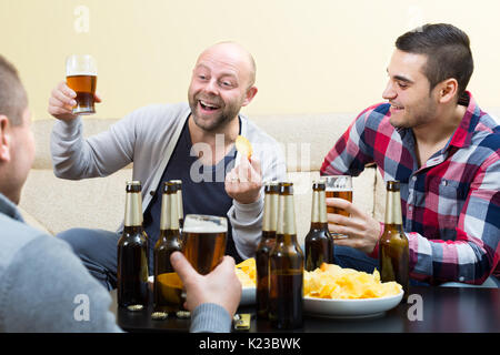 Three happy friends drinking beer  with crisps and talking at home Stock Photo