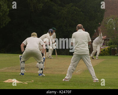 Close view village cricket match on the village green at Benenden in Kent with batsman, wicket keeper and close fielders and ball leaving bowlers hand
