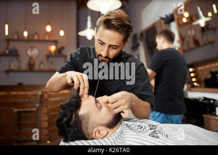 Hairdresser working at barbershop, correcting bread with white thread. Stock Photo