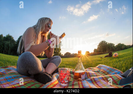 A young girl enjoys a lollipop and a glass of wine on a rug in summer whilst texting on her cellphone Stock Photo