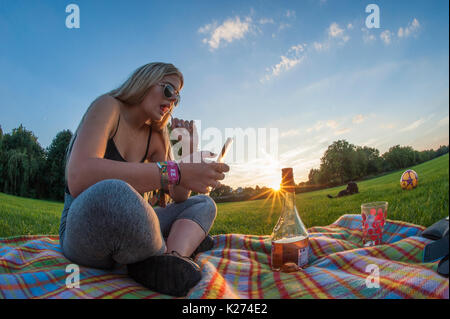 A young girl enjoys a lollipop and a glass of wine on a rug in summer whilst texting on her cellphone Stock Photo