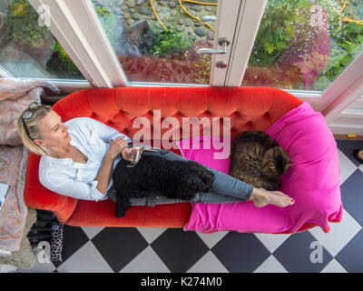 A woman reclines on a bright red sofa with her dog and cat on her lap, looking at her cell phone Stock Photo