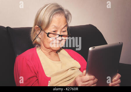 Old woman wearing glasses seated on a sofa at home reading something on a touchscreen tablet. Stock Photo