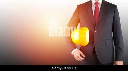 Engineer in black suit holding yellow helmet on gray background with sunlight Stock Photo