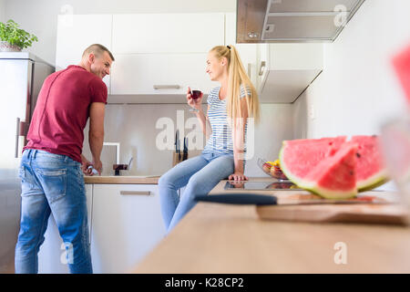 Attractive young caucasian man washing up dishes, while his girlfriend who is sitting on kitchen counter, holds glass of red wine Stock Photo