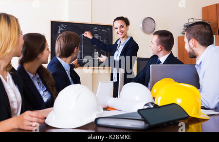 Portrait of professionals with helmets and laptops having meeting in office interior. Focus on standing woman Stock Photo
