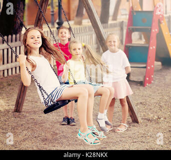 Laughing girls and boys in school age swinging on children's playground outdoors Stock Photo