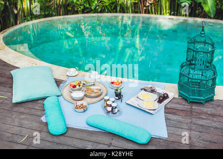 Cozy organic breakfast on plaid for two near private pool. Vintage style, all natural and homemade: muesli, jam, bread, yogurt, scrambled eggs, fresh  Stock Photo