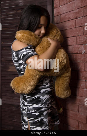 Sad little girl embracing her teddy bear. punished girl standing near brick wall Stock Photo