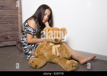 Sad little girl embracing her teddy bear. punished girl sitting near white wall Stock Photo