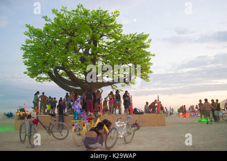 Burners gather around a giant tree sculpture at sunset on the playa as the annual desert festival Burning Man closes the first day of the week-long event August 27, 2017 in Black Rock City, Nevada. The annual festival attracts 70,000 attendees in one of the most remote and inhospitable deserts in America. Stock Photo