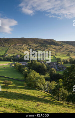 High view over beautiful, sunny Yorkshire Dales village of Burnsall in valley by River Wharfe & with steep, green, hillside beyond - England, GB, UK.